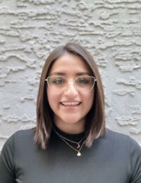 A woman with straight brown hair, glasses, and a black top stands in front of a textured light gray wall, smiling at the camera. She wears layered gold necklaces, reflecting the warm spirit behind our Acerca de nosotros story. - Mereces Jeremías Stefany Gomez