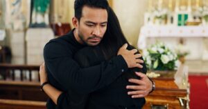 A man and woman dressed in black embrace in a church, standing beside a wooden casket with flowers in the background.
