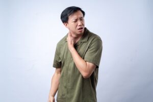 A man in a green shirt touches his neck and appears to be in discomfort, standing against a plain light background.