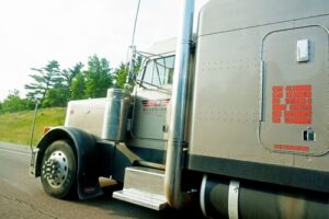 A silver semi truck driving on a road with trees and grass in the background. - Mereces Jeremías A silver semi truck driving on a road with trees and grass in the background.