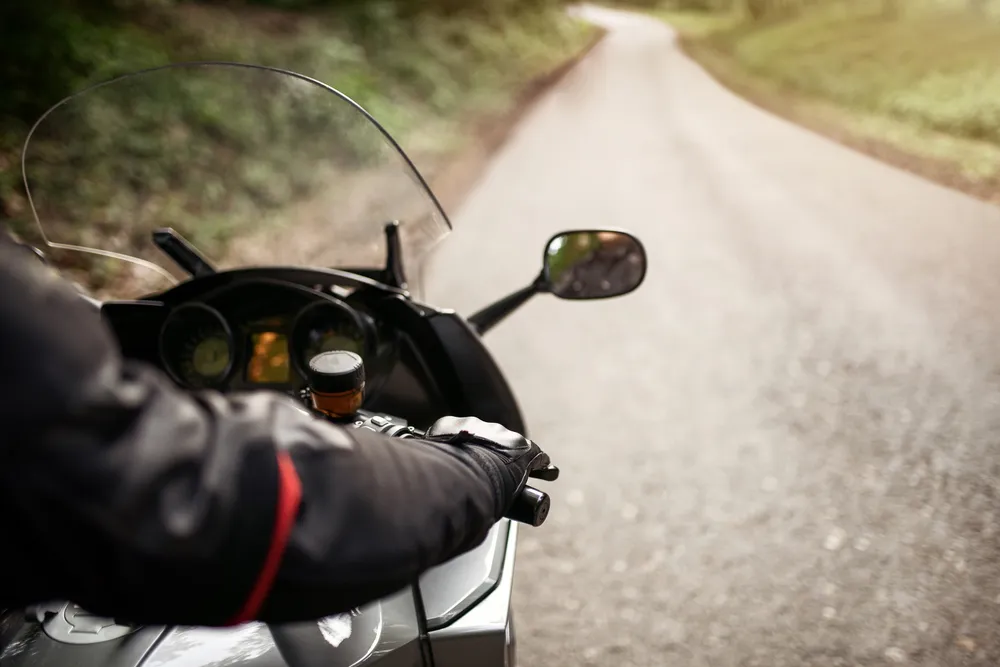 A motorcyclist wearing a black jacket rides a motorcycle on an empty, winding road bordered by greenery.