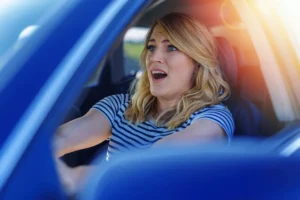 A woman with blonde hair wearing a striped shirt sits in the driver's seat of a car, looking surprised or alarmed, with her mouth open and hands on the steering wheel. A woman with blonde hair wearing a striped shirt sits in the driver's seat of a car, looking surprised or alarmed, with her mouth open and hands on the steering wheel.