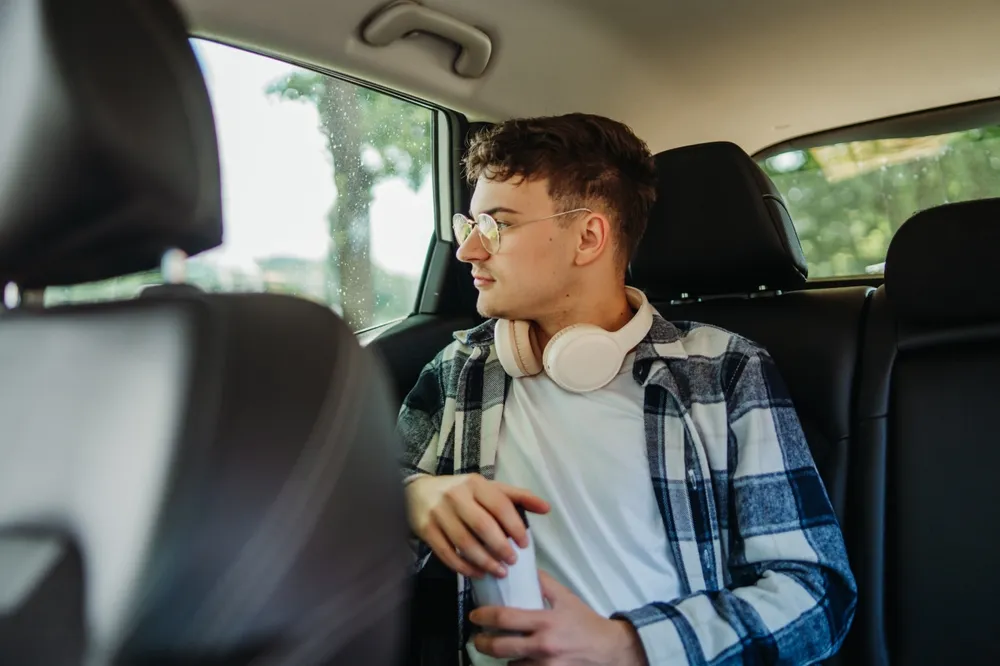Young man with glasses and headphones around his neck sits in the back seat of a car, looking out the window.