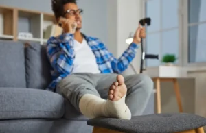 A person with a bandaged foot is sitting on a couch, talking on the phone, and holding crutches, with their injured leg resting on a stool.