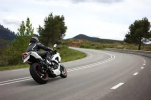 A motorcyclist in black gear rides a white sport bike on a winding, empty road with trees and hills in the background.