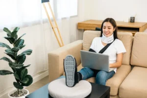 A woman with a neck brace and arm sling sits on a couch with her leg elevated, using a laptop. Crutches are propped against the wall nearby.