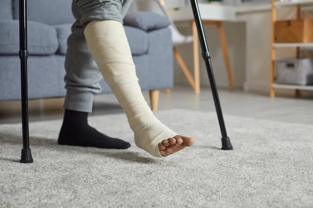 Person with a leg cast and crutches stands on a carpeted floor in a living room. Only the lower body is visible.