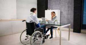 A person in a wheelchair with arm and neck braces sits across a desk from a woman using a laptop in an office setting. - Mereces Jeremías A person in a wheelchair with arm and neck braces sits across a desk from a woman using a laptop in an office setting.