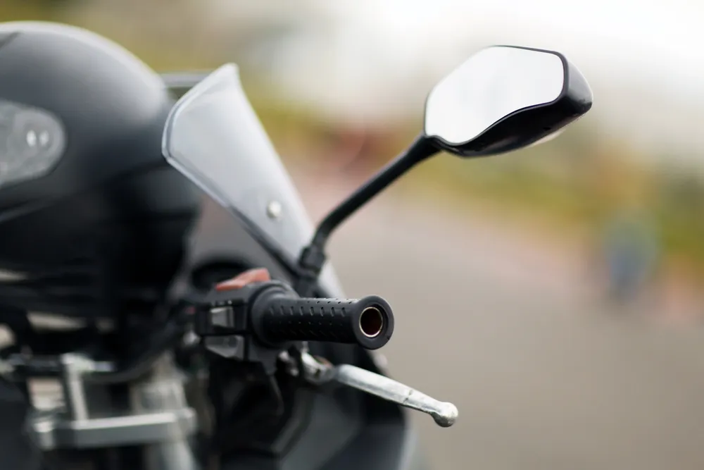 Close up of a motorcycle handlebar with a rearview mirror and brake lever, with a blurred background.