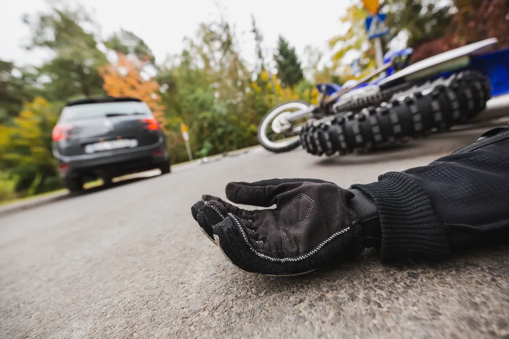 A gloved hand lies on the road near a fallen motorcycle, with a car parked nearby and trees in the background, suggesting an accident scene.