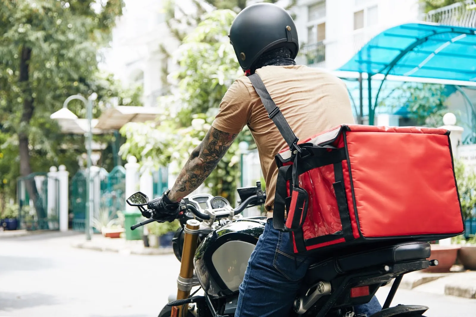 A motorbike delivery rider wearing a helmet and carrying a large red insulated food bag is stopped outdoors in a residential area.
