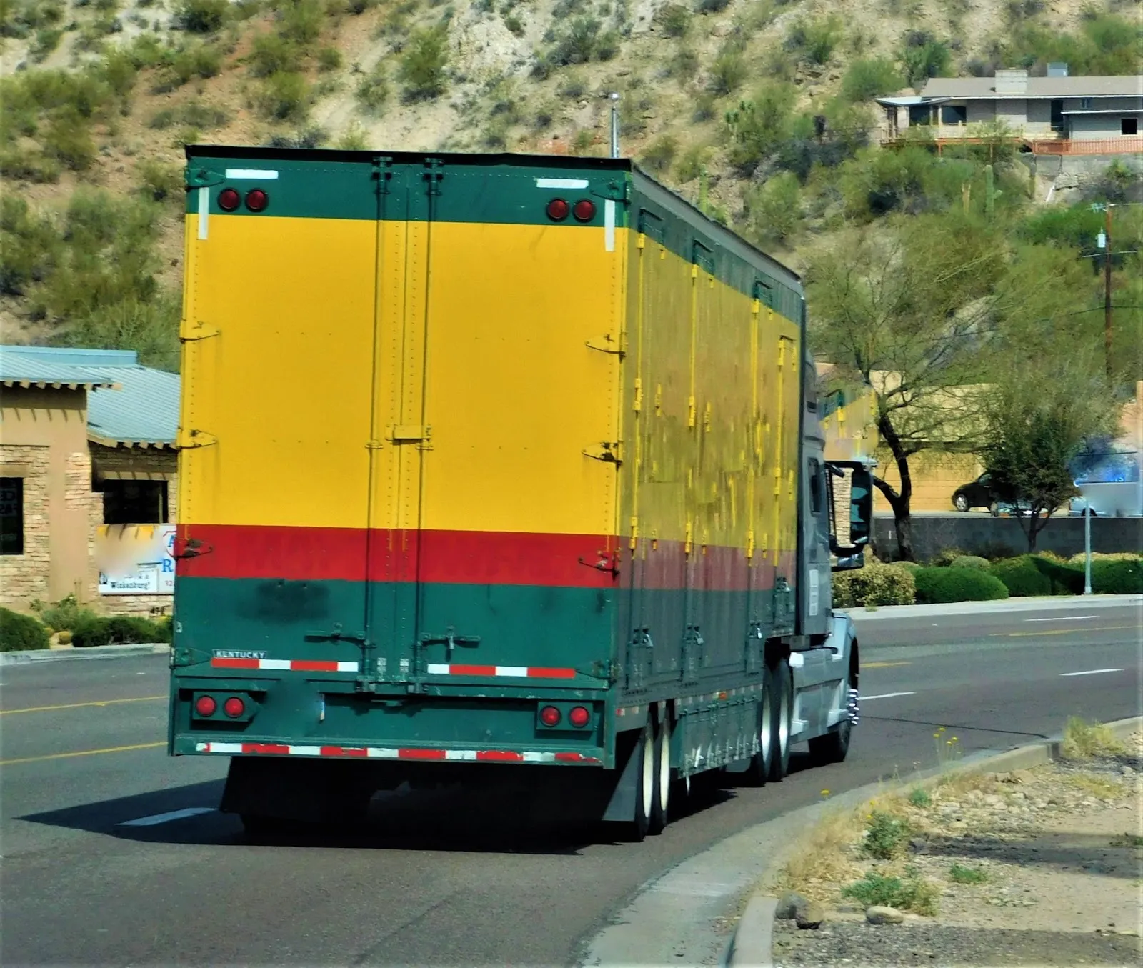 A large green and yellow semi truck drives on a curved road near buildings and a hillside with scattered vegetation.