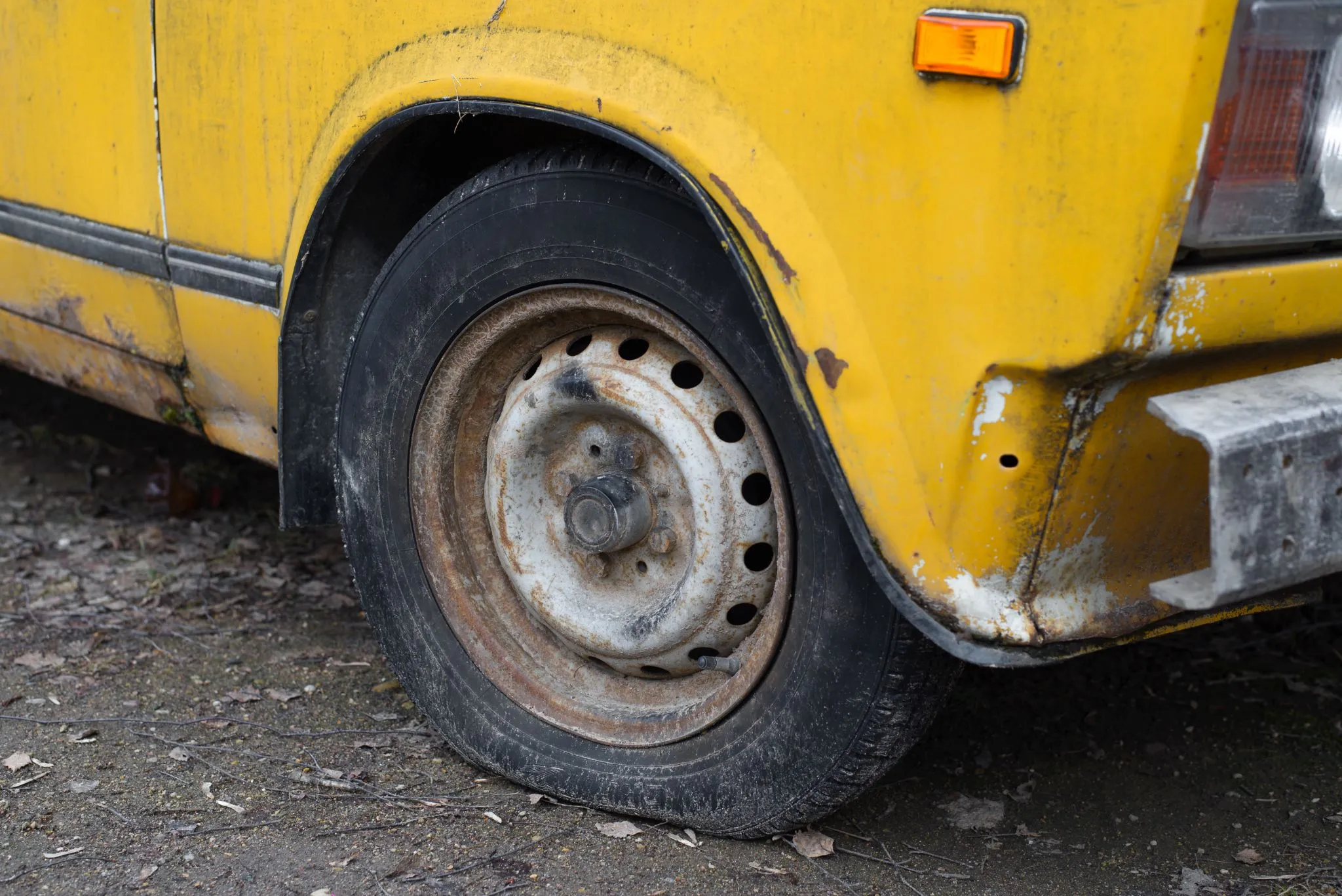 Close up of a flat front tire on an old, yellow car with visible rust and dirt on the body, parked on a rough, leaf covered ground.