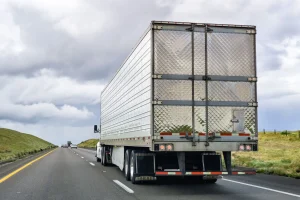 A large silver semi truck drives on a highway with grassy hills on both sides under a cloudy sky.