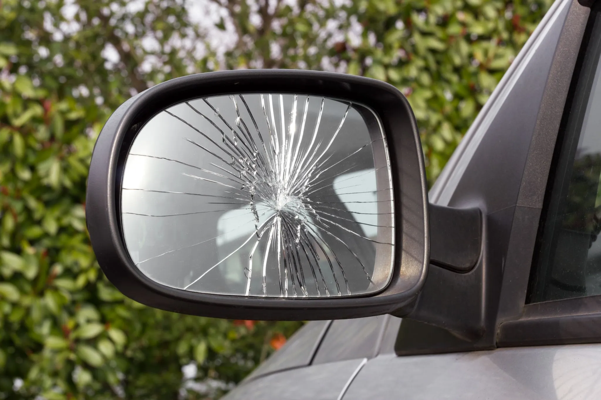 A car’s side mirror with a large crack running across the glass, reflecting the surrounding greenery.