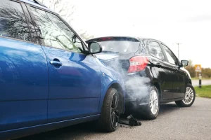 A blue car has rear ended a black car on a road, causing visible damage and smoke between the vehicles. A blue car has rear ended a black car on a road, causing visible damage and smoke between the vehicles.