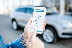 Person holding a smartphone displaying a navigation app with a mapped route, standing outdoors near a parked silver SUV. - Mereces Jeremías Person holding a smartphone displaying a navigation app with a mapped route, standing outdoors near a parked silver SUV.
