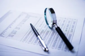 A magnifying glass and a pen rest on a printed spreadsheet with rows and columns of numbers. - Mereces Jeremías A magnifying glass and a pen rest on a printed spreadsheet with rows and columns of numbers.