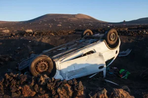 A white car lies overturned on rocky ground in a barren landscape with hills in the background.