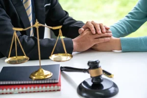 A person in a suit comforts another by holding their hands at a desk with legal scales, a gavel, and notebooks.