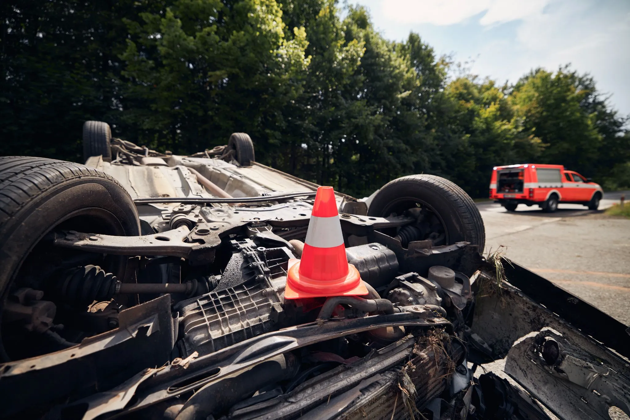 A flipped car lies upside down on the road with a traffic cone placed on its undercarriage; an emergency vehicle is parked in the background near some trees.