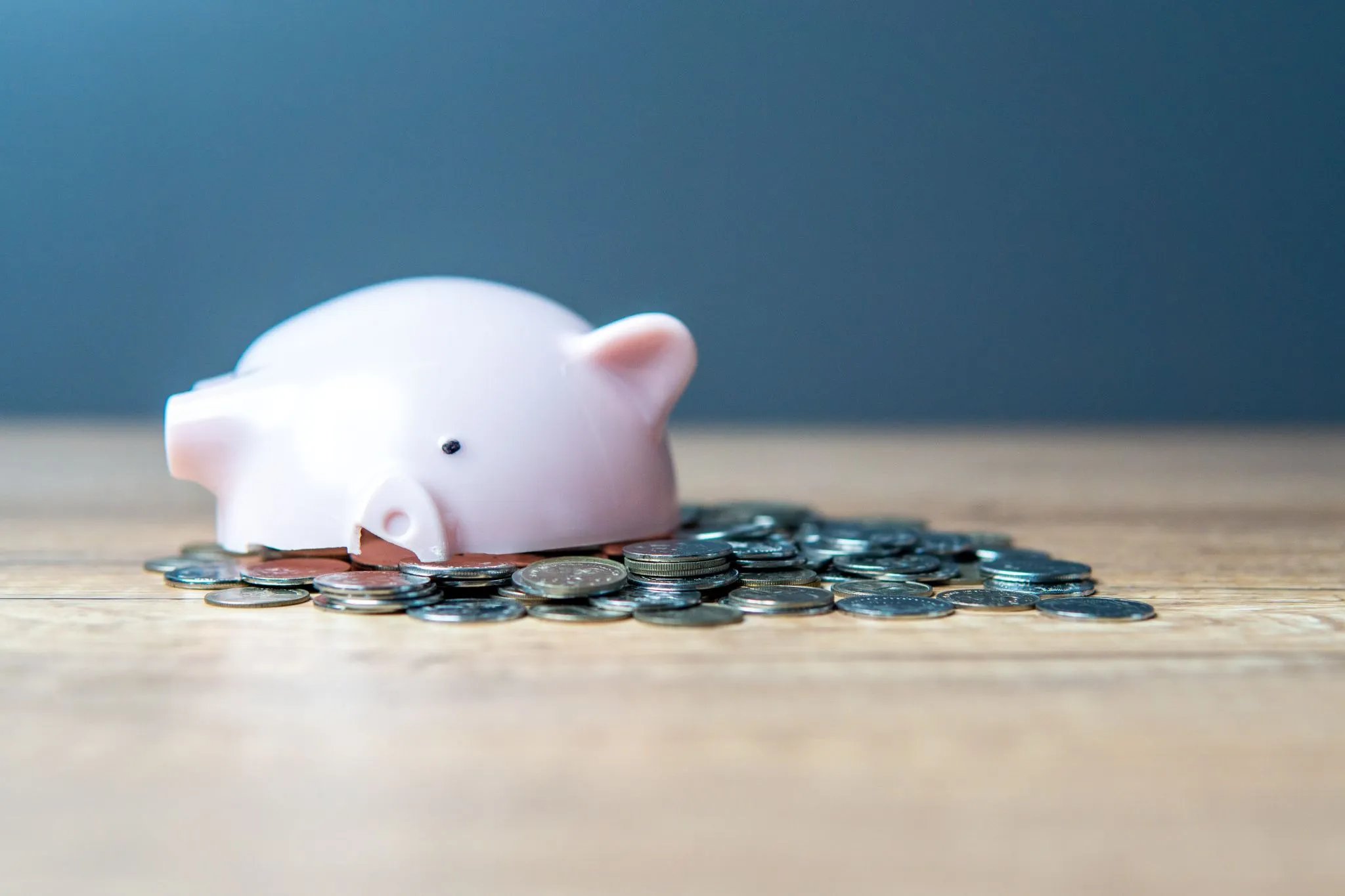 A pink piggy bank lying on its side with coins scattered around it on a wooden surface.