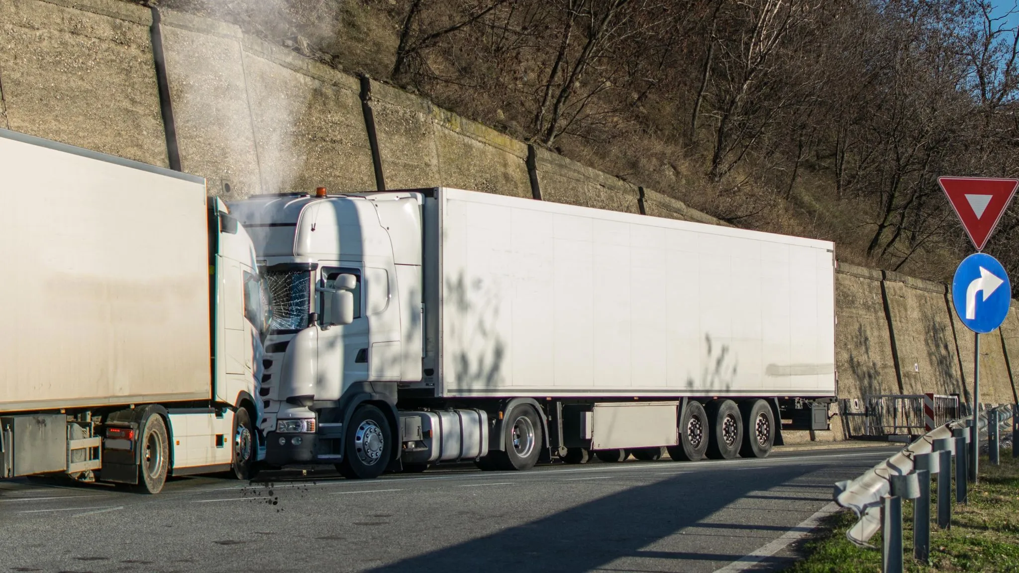 Two large white semi trucks have collided front to front on a road near a stone retaining wall, with visible damage and steam coming from one truck.