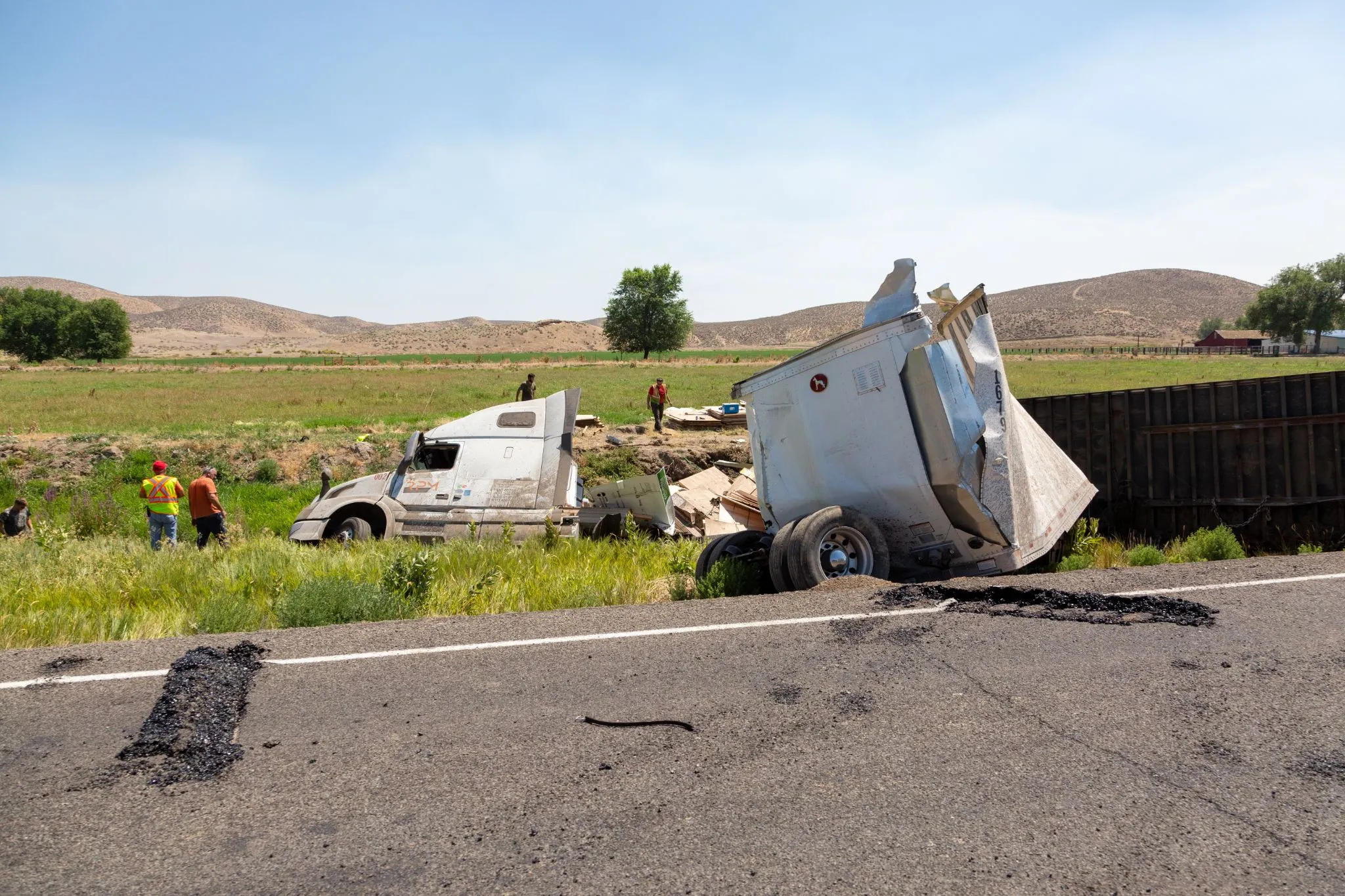 A jackknifed semi truck lies in a ditch beside a rural road as workers in safety vests assess the accident scene under a clear sky.