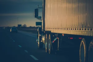 A large semi truck travels down a highway at dusk, with cars visible in adjacent lanes and the sun casting a golden reflection on the truck - Mereces Jeremías's trailer. A large semi truck travels down a highway at dusk, with cars visible in adjacent lanes and the sun casting a golden reflection on the truck's trailer.