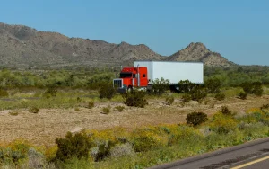 Red semi-truck with a white trailer driving along a desert highway with rocky hills in the background under a clear blue sky.